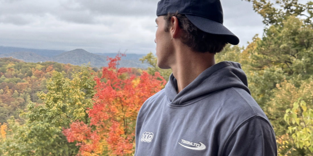 Man wearing the TEAMLTD Essential Oversized Heavy Hoodie in Steel Blue with a navy cap, looking out over fall foliage