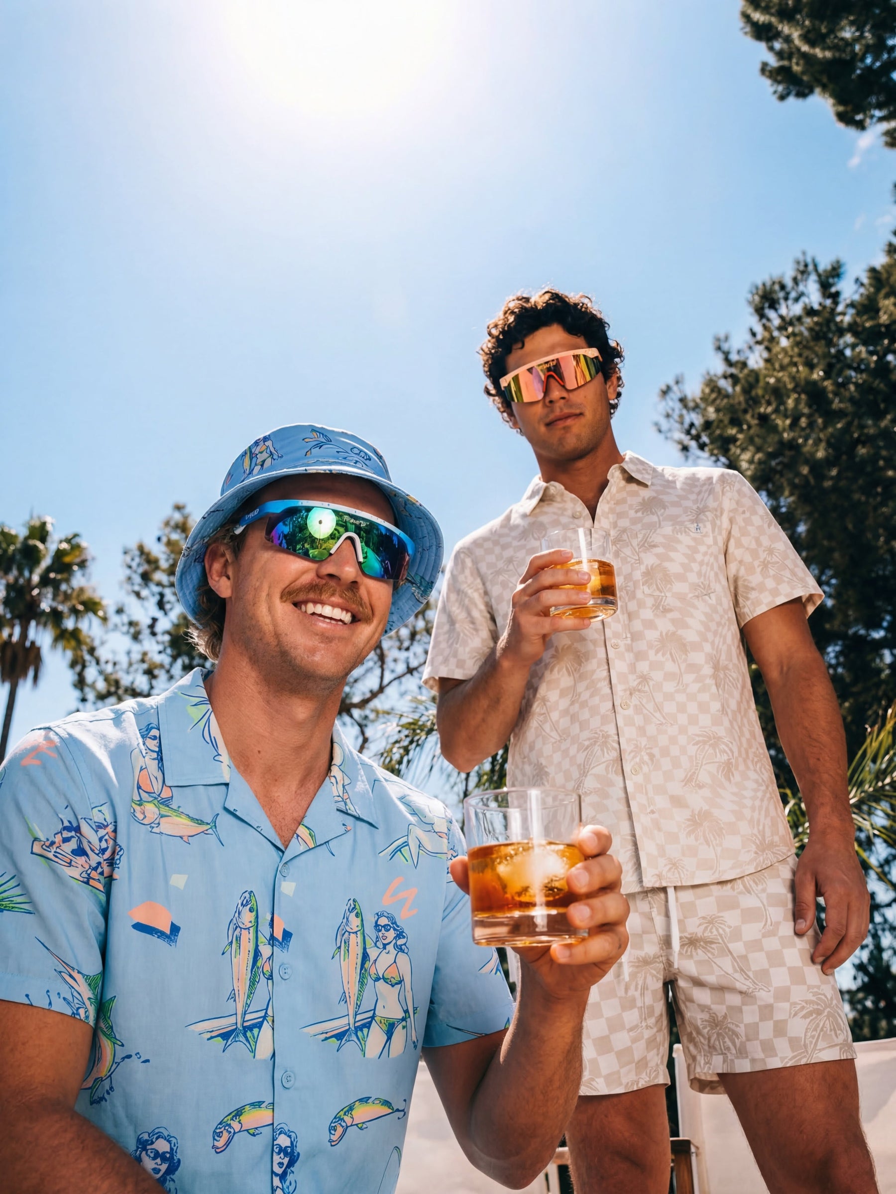 Two men outdoors under a bright sun wearing matching tropical resort outfits. The man in the foreground wears a light blue bucket hat and a light blue short-sleeve button-up shirt with a multicolor fish and turtle print, holding an iced drink.